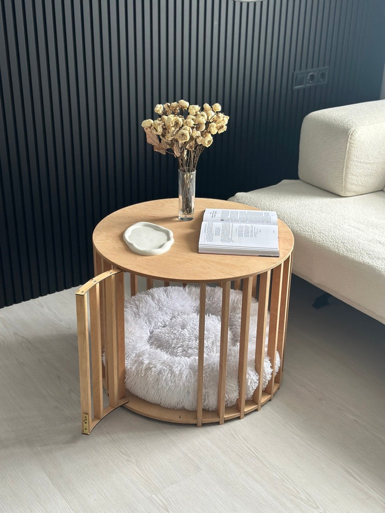 Round wooden side table with pet bed, vase of flowers, and book in a living room setting.